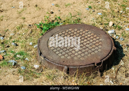 Cast iron cover on the hatch of a sewer well among grass and sand. Stock Photo