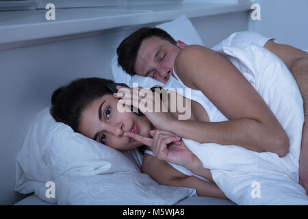Young Woman Talking On Mobile Phone While Her Husband Sleeps On Bed Stock Photo