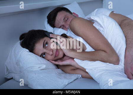 Young Woman Using Mobile Phone On Bed While Her Husband Sleeping Stock Photo