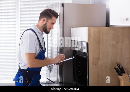 Young Male Electrician In Overall Writing On Clipboard In Front Of Oven Stock Photo