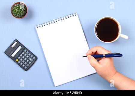 Empty notebook with pen, coffee, calculator and cactus on a blue background. Copy the space. Top view. The hand holds a pen Stock Photo