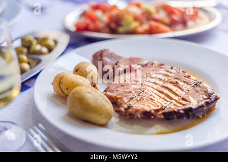 Tuna steak accompanied with potatoes, olives, tomato salad and wine on white plate Stock Photo
