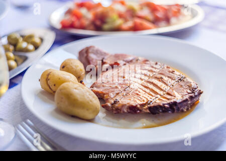 Tuna steak accompanied with potatoes, olives, tomato salad and wine on white plate Stock Photo