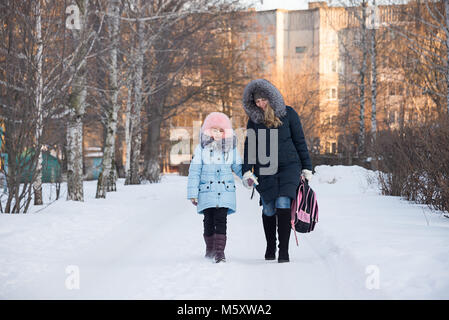 a young mother leads her daughter from school, the family smiles, children have fun, the family walks in the winter, go on the road. Stock Photo