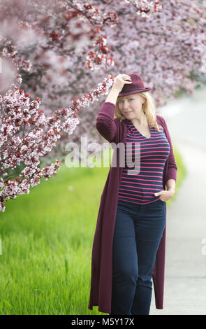 Confident Caucasian woman with long blonde hair near blossoming orchard with purple fedora hat Stock Photo