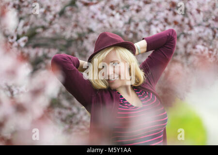 Caucasian woman with long blond hair and purple fedora hat near blossoming tree, arms behind her head with the elbows pointing out Stock Photo