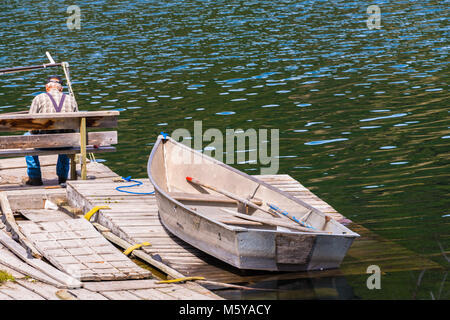 Old Wooden Row Boat on Dock with Old Man Sitting on a Bench and Fishing . Copy space. Stock Photo