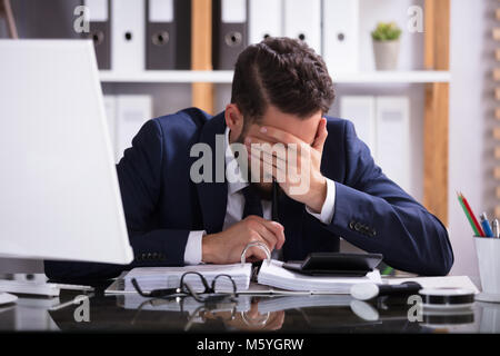 Young Man Suffering From Headache Working In Office With Bill Over Desk Stock Photo