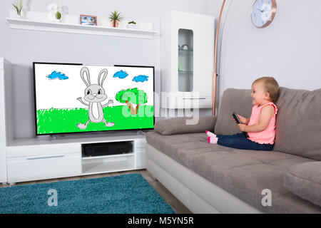 Happy Baby Girl Sitting On Sofa Watching Cartoon On Television Stock Photo