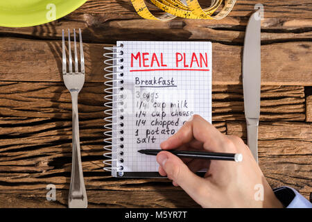 Elevated View Of Hand Making Meal Plan On Notebook With Fork And Knife On Desk Stock Photo