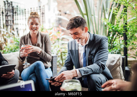 Businessman is enjoying drinks in a bar courtyard with colleagues. He is laughing while using his smart phone. Stock Photo