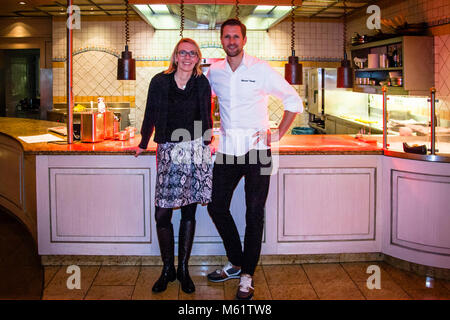 German two-star chef Michael Kempf stands with reporter Angela Berg in front of the open kitchen at the Parkhotel Ahrensburg, Germany. His white chef's jacket carries a visible name tag reading "Michael Kempf," with the red counters and shelves of the open show kitchen visible behind them. Kempf is head chef of the two-Michelin-star Berlin restaurant Facil at Hotel Mandala, identifiable from the embroidered "Facil" lettering on his brown apron in another photo from the same evening. Stock Photo