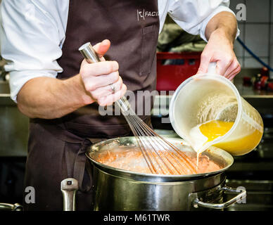 German two-star chef Michael Kempf pours a creamy blood-orange hollandaise over a simmering pot in the kitchen of the Parkhotel Ahrensburg, Germany, while stirring with a whisk in his other hand. His wedding ring is clearly visible on his right hand, and his brown apron carries the embroidered "Facil" lettering. The sauce is part of one of the courses Kempf, head chef of Berlin's two-Michelin-star restaurant Facil, prepares that evening. Stock Photo