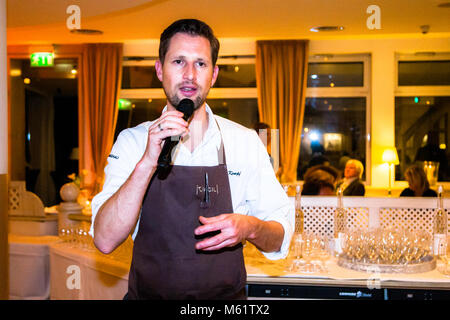 German two-star chef Michael Kempf speaks with a microphone in front of guests in the festively lit dining room of the Parkhotel Ahrensburg, Germany. His brown apron carries an embroidered name, with orange curtains and a tray lined with champagne glasses visible in the background. Kempf, head chef of Berlin's two-Michelin-star restaurant Facil, appears to be introducing one of the served courses to the guests. Stock Photo