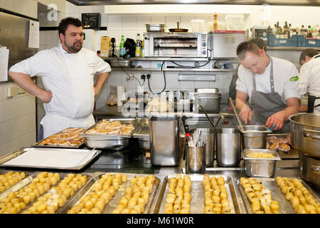 German Michelin-starred chef Sebastian Prüßmann prepares his potato lunch in the kitchen of the Hotel and Restaurant Kronenschlösschen in Hattenheim, Eltville am Rhein, Hesse, Germany. He stands with his hands on his hips in front of trays full of peeled potatoes. On the right a young cook in a grey apron works with a knife at a stainless steel container. Trays of seared meat rolls stand behind them. At the South Africa lunch of the 21st Rheingau Gourmet & Wein Festival on 12 March 2017 Prüßmann cooks four courses: marinated black salsify with Périgord truffle, pan-fried monkfish with cuttlefi Stock Photo