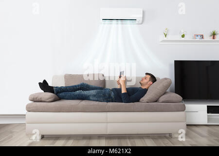 Young Man Lying Under Air Conditioner On Couch Using Tablet At Home Stock Photo