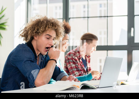 Funny student yawning in front of a book while sitting down at desk Stock Photo
