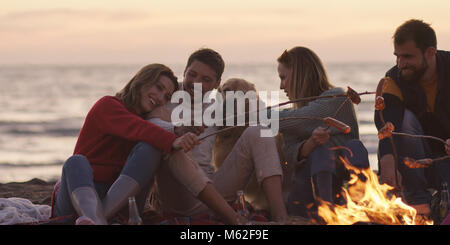 Group of young friends sitting by the fire late at night, grilling sausages and drinking beer, talking and having fun Stock Photo