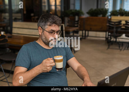 Working online. Freelancer man with beard and eyeglasses working in coffee shop using black laptop with caramel macchiato by his side. Stock Photo