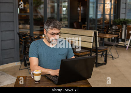 Working online. Freelancer man with beard and eyeglasses working in coffee shop using black laptop with caramel macchiato by his side. Stock Photo