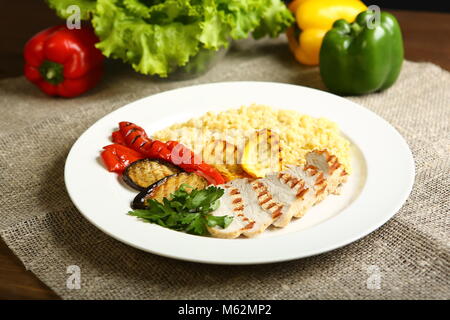 Grilled chicken with slices, with vegetables, bulgur on a white plate, decorated with parsley. In the background, lettuce leaves, color pepper. Low-ca Stock Photo
