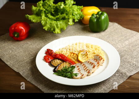 Grilled chicken with slices, with vegetables, bulgur on a white plate, decorated with parsley. In the background, lettuce leaves, color pepper. Low-ca Stock Photo
