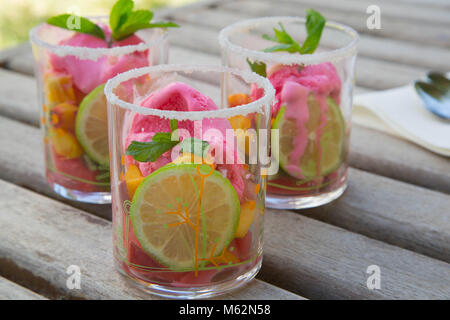 Three glasses with fruit salad and strawberry ice cream on the wooden surface. Spoons in the background Stock Photo