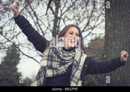 Joyful brunette girl smiling with arms up in the park on cloudy autumn season day. Celebration concept. Pretty young woman happy Stock Photo