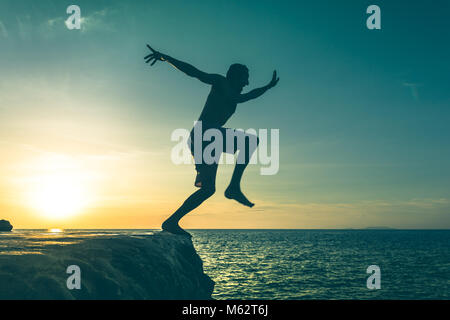 Man jumping over a cliff into the sea on sunset in Koh Phangan island, Thailand. Vintage effect. Dare, fearless concept. Two out of three series Stock Photo