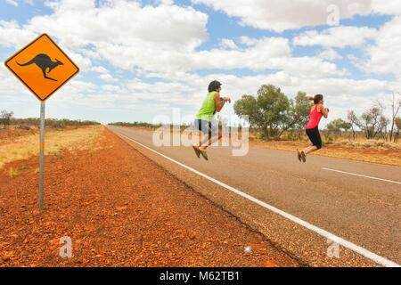 Funny couple of tourists pretend jumping like kangaroos by road sign on traditional empty highway in Northern Territory, Outback Australia Stock Photo