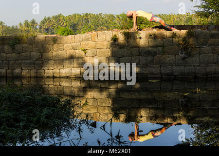 Yogi woman in purvottanasana pose on top of rocks wall with reflection on river by Srirangapatna, Karnataka, India. Nature connection concept Stock Photo