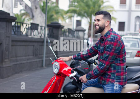Young hipster with smile sitting on red scooter in the city. Handsome full bearded man parking motorcycle on street Stock Photo