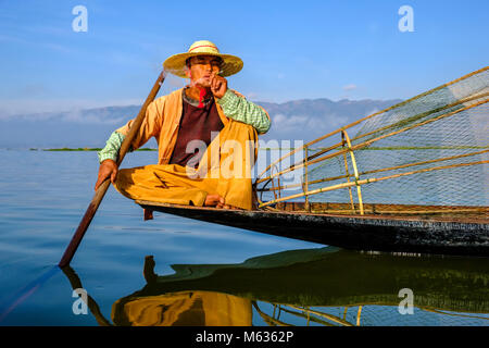 A fisherman, sitting on his boat smoking, is fishing the traditional way on Inle Lake Stock Photo