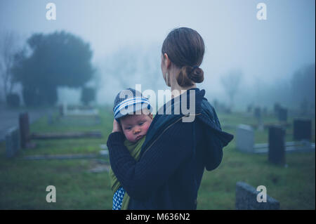 A grieving young mother is standing with her baby by a tombstone in a graveyard Stock Photo