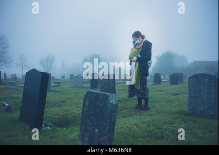 A grieving young mother is standing with her baby by a tombstone in a graveyard Stock Photo