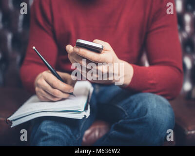 A man is sitting on a sofa using a smartphone and writing in a notebook Stock Photo