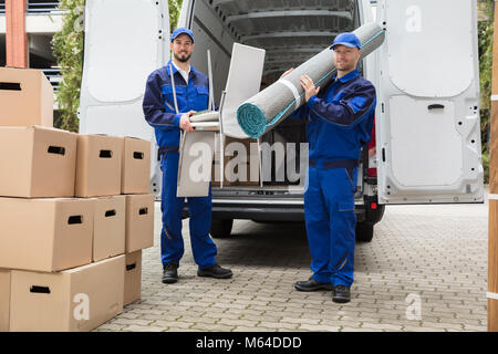 Two Smiling Delivery Men Holding Chairs And Carpet In Front Of Truck Stock Photo