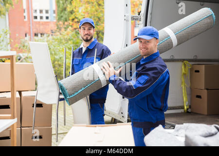 Two Smiling Delivery Men Holding Chairs And Carpet In Front Of Truck Stock Photo