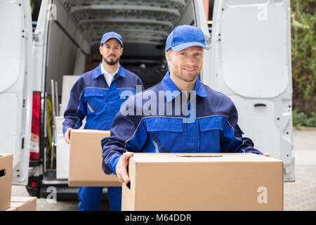 Close-up Of Two Young Delivery Men Carrying Cardboard Box In Front Of Truck Stock Photo