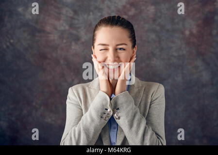 Cute attractive young woman winking as she grins happily at the camera while holding her hands to her cheeks over a textured slate background Stock Photo