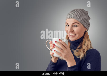 Smiling happy blond woman wearing a knitted winter hat and polo neck sweater standing indoors with a large mug of hot coffee in her hands looking at t Stock Photo