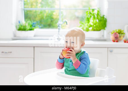 Baby eating fruit. Little boy biting apple sitting in white high chair in sunny kitchen with window and sink. Healthy nutrition for kids. Solid food f Stock Photo
