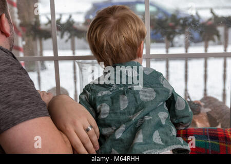 A Young Caucasian Father and Son Sit On Their Living Room Couch Looking Out The Window At Falling Snow Stock Photo