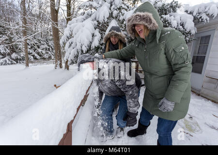 Two Yong Women In Winter Coats Playfully Push A Young Mans Face Into The Snow Stock Photo