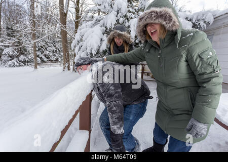 Two Yong Women In Winter Coats Playfully Push A Young Mans Face Into The Snow Stock Photo