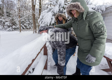 Two Yong Women In Winter Coats Playfully Push A Young Mans Face Into The Snow Stock Photo