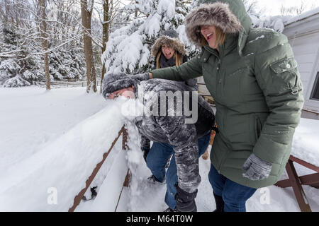 Two Yong Women In Winter Coats Playfully Push A Young Mans Face Into The Snow Stock Photo