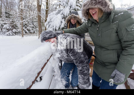 Two Yong Women In Winter Coats Playfully Push A Young Mans Face Into The Snow Stock Photo