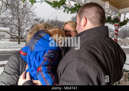 Two Caucasian Women And A Young Toddler Boy Stand On A Front Porch As It Snows Looking At The Christmas Decorations. Stock Photo