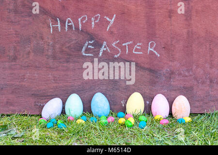 Colorful Easter eggs on grass and rugged wooden background with happy Easter written with chalk. Traditional hand-painted eggs for Easter celebration Stock Photo